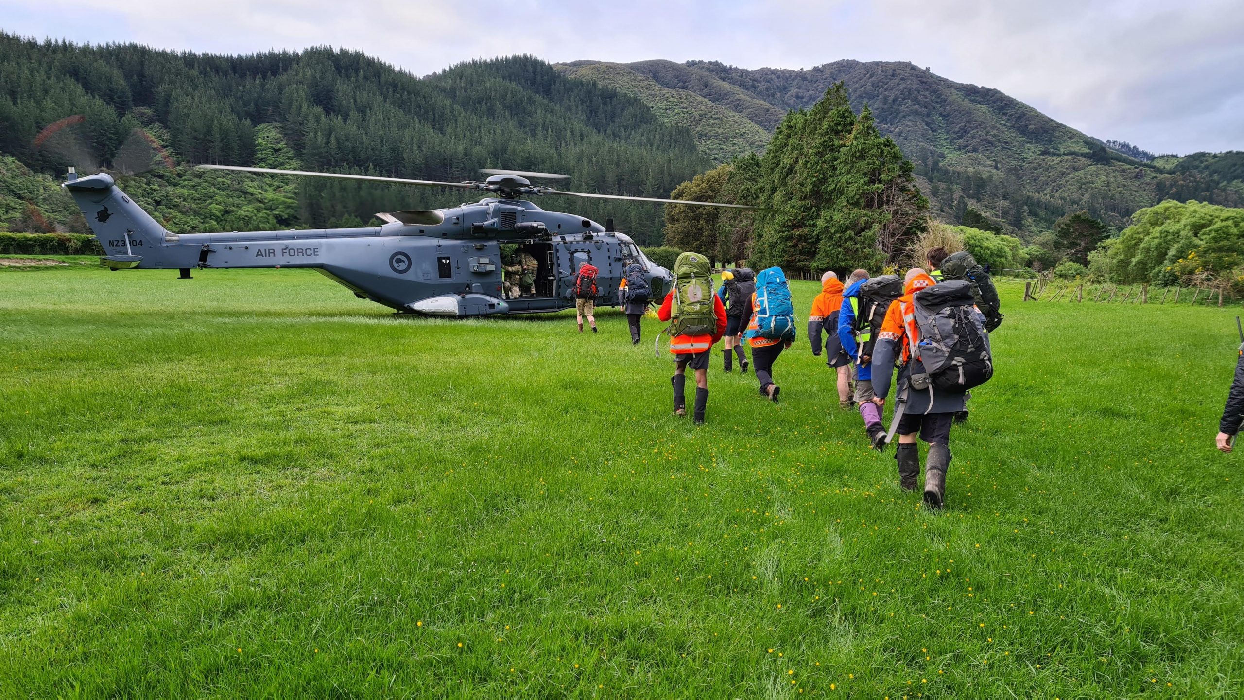 Wellington Land Search and Rescue Members with packs and equipment walking towards an NH90 helicopter that has landed in a large green field.