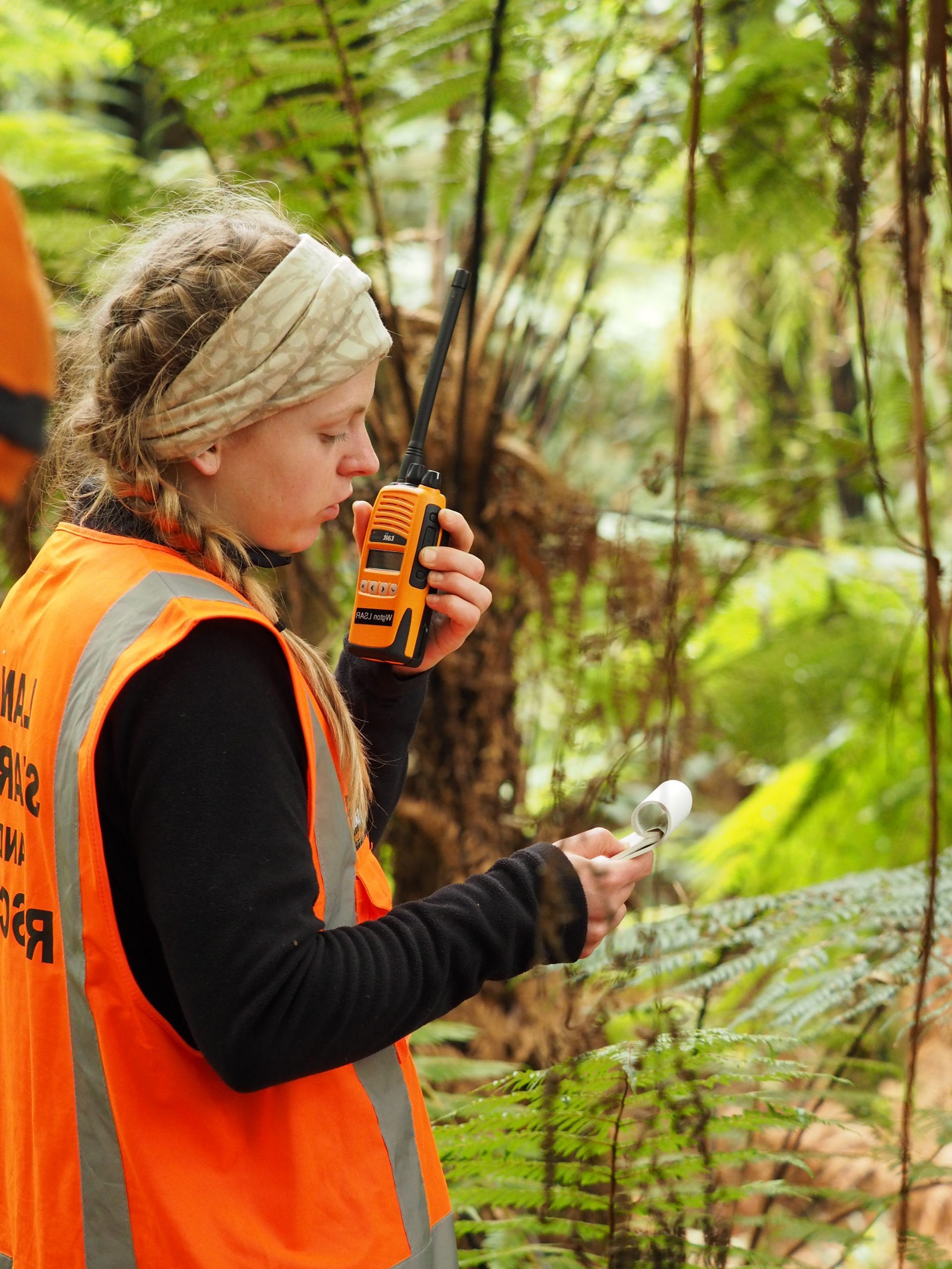 Female LandSAR volunteer in the bush. She is wearing an orange LandSAR hi-vis vest, and is reading from her notebook while transmitting via an orange handheld radio