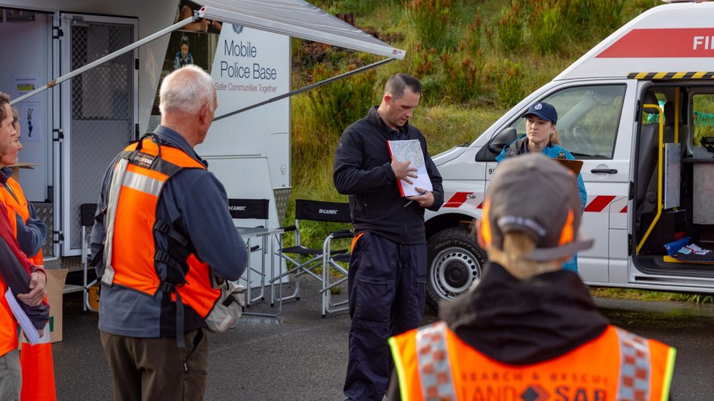 A male Police officer stands in front of a group of LandSAR volunteers in their orange hi-vis uniforms. He is pointing to a map and briefing the volunteers.