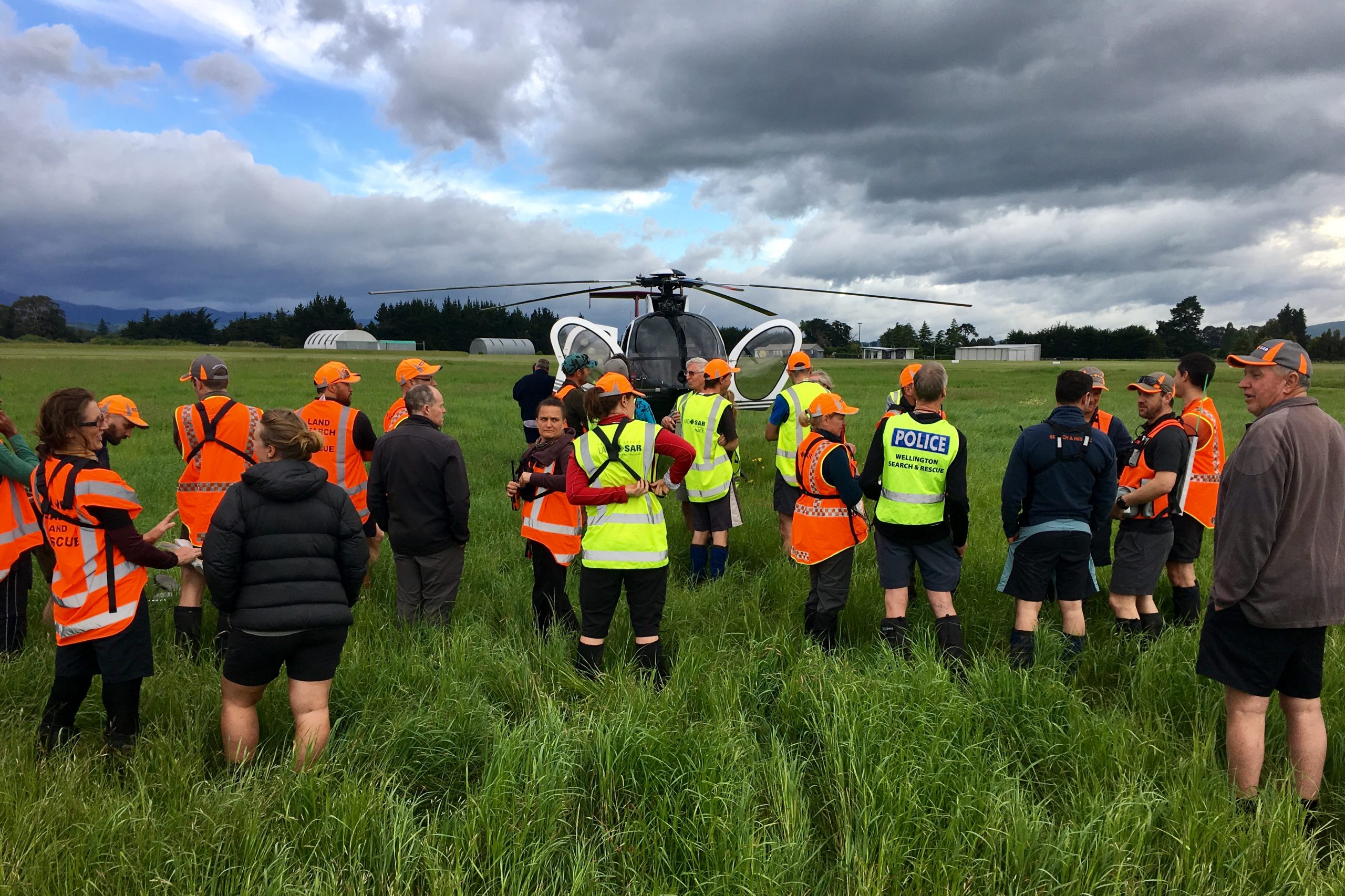 A large group of LandSAR volunteers stand in an open field. They are wearing their hi-vis uniforms, and standing in front of a helicopter.