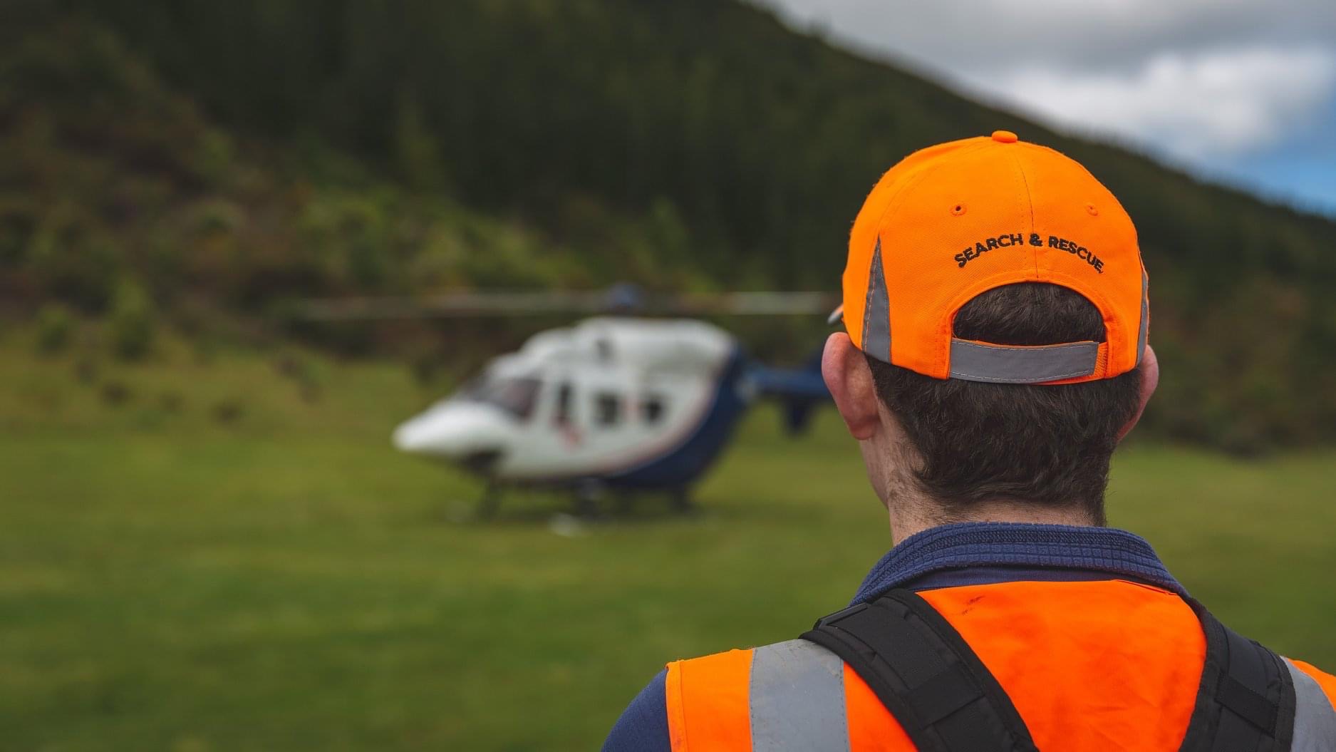 Man wearing hi-vis LandSAR cap and vest. He is standing in front of a rescue helicopter.