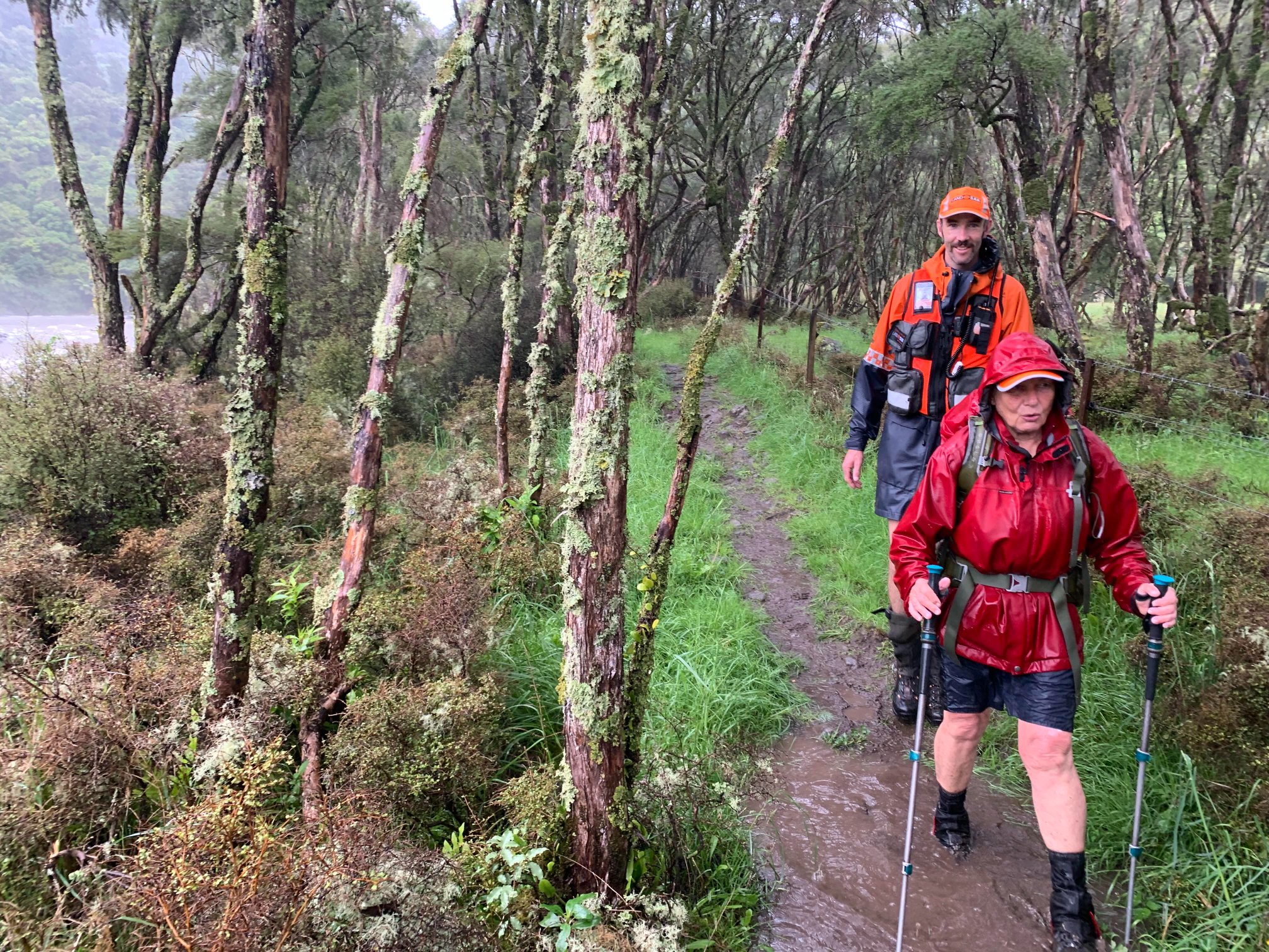 A woman and a man wearing rain jackets tramp along a track through the trees in the rain.