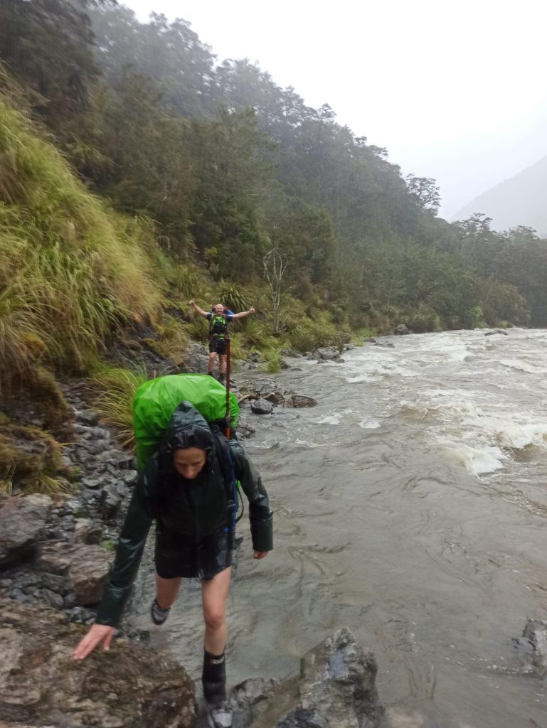 Woman searcher wearing rain jacket and carrying a green pack. She is clambering along a swollen river.