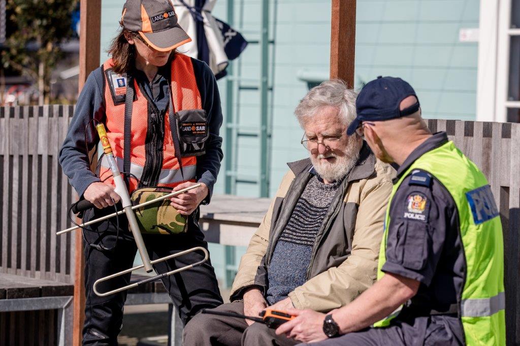 Elderly man sitting on a bench, appearing confused. He is sitting next to a Police Officer wearing blue overalls and a hi-vis vest. Standing next to both of them is a woman wearing an orange Land Search and Rescue hi-vis vest. She is holding a large aerial that is used for searching for pendants worn by WanderSearch users.